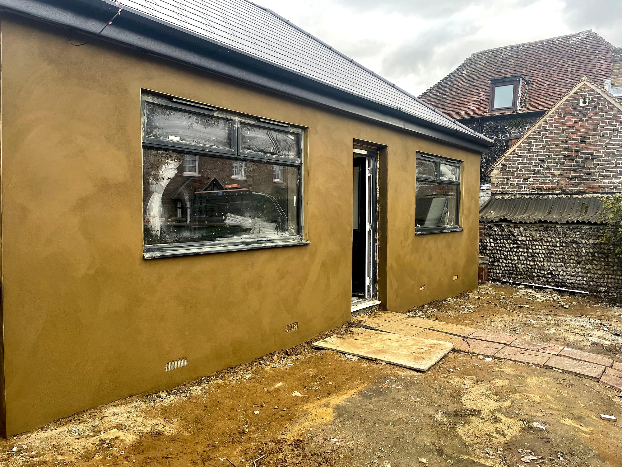 Newly rendered single-storey extension with ochre sand and cement finish beside traditional flint wall in East Kent