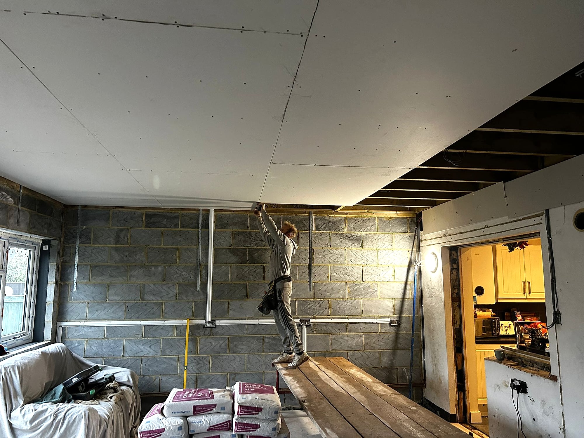 Lee Wehrle fitting plasterboard ceiling in blockwork extension standing on scaffold platform in Kent