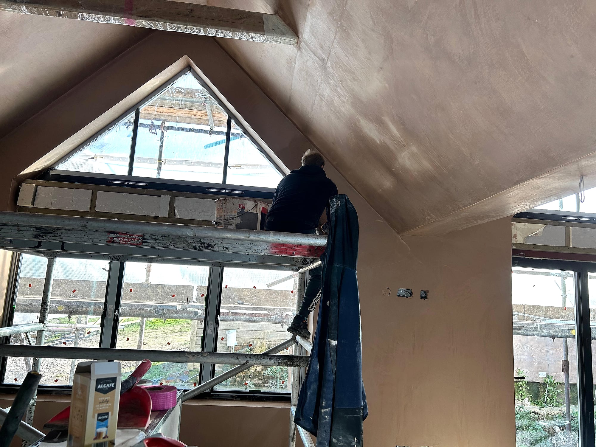 Lee plastering a vaulted ceiling from scaffolding platform beside triangular apex window in Kent extension
