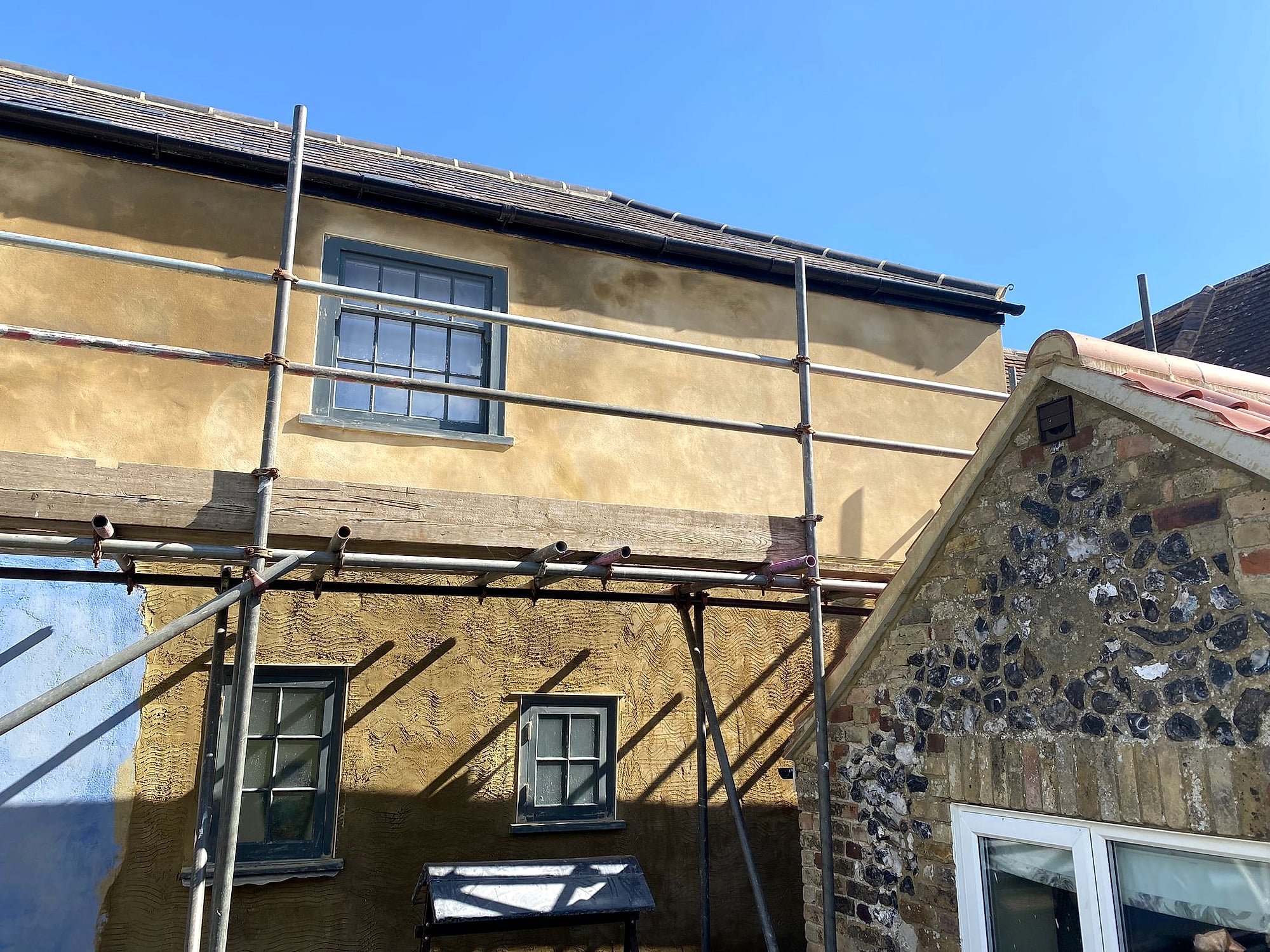Period cottage with completed sand and cement render next to flint outbuilding against blue sky in East Kent