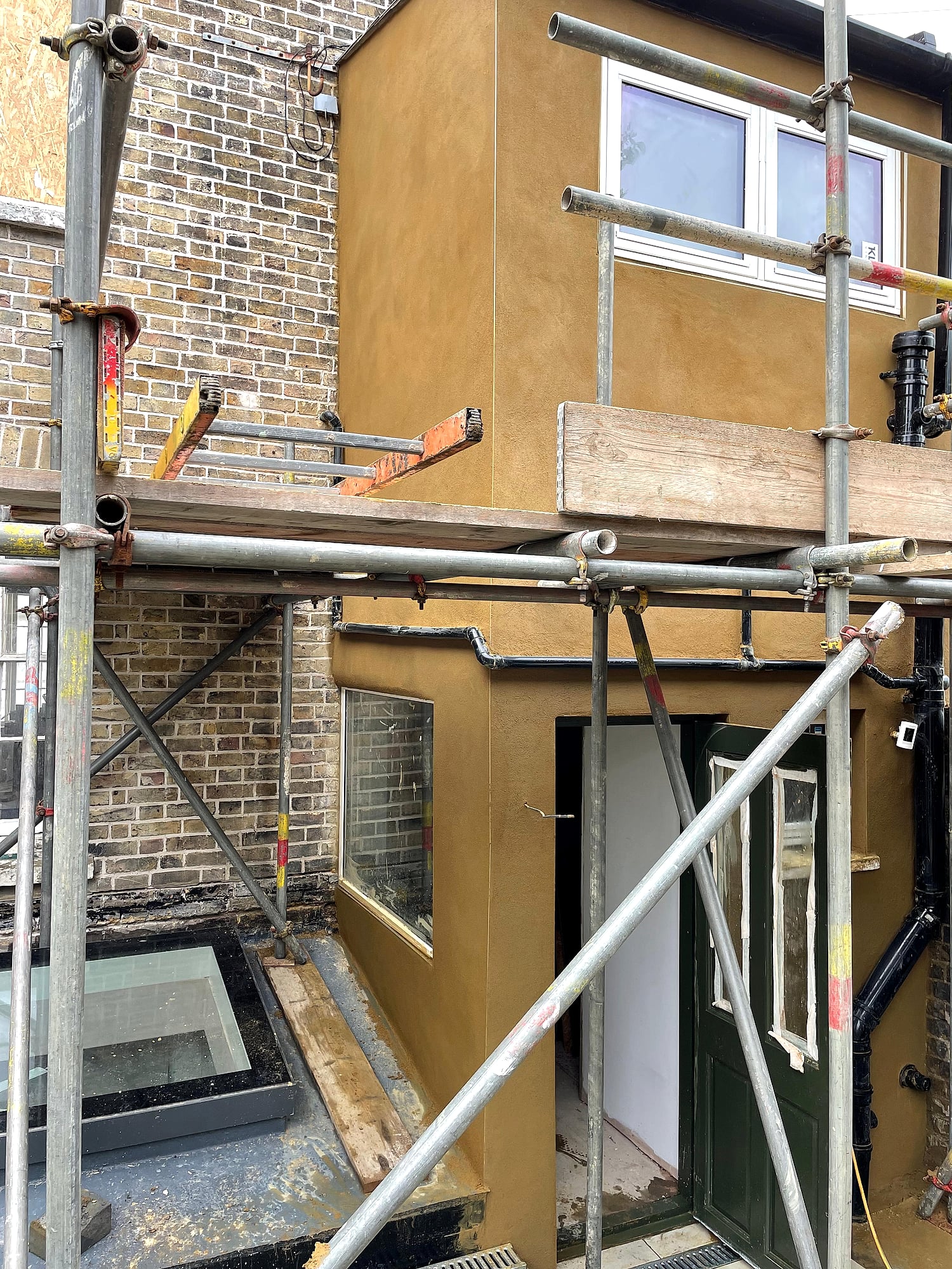 Rear extension with fresh ochre sand and cement render applied from scaffolding next to brick wall in East Kent