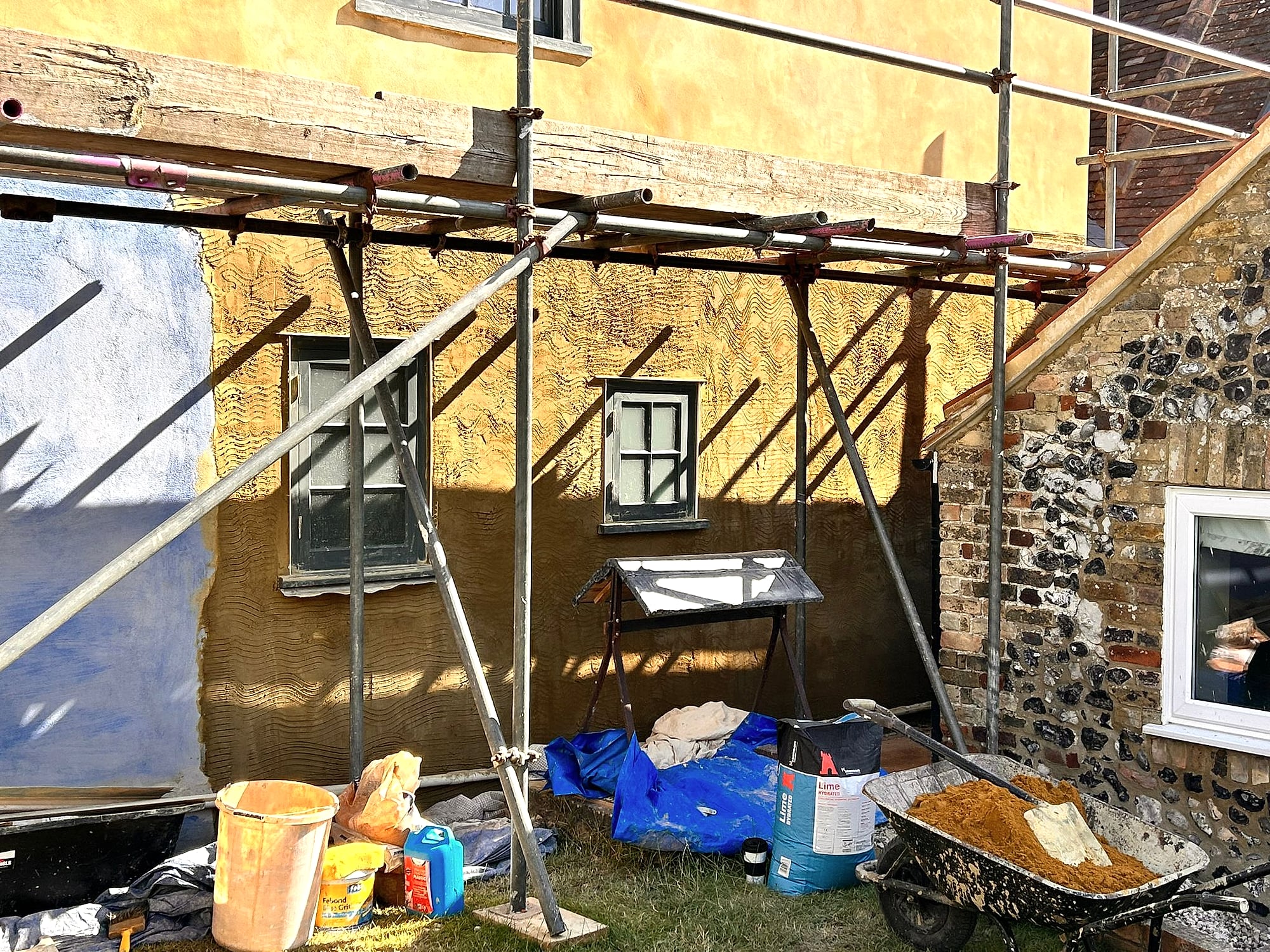 Scratch coat render being applied to period cottage wall next to flint outbuilding with scaffolding in Kent